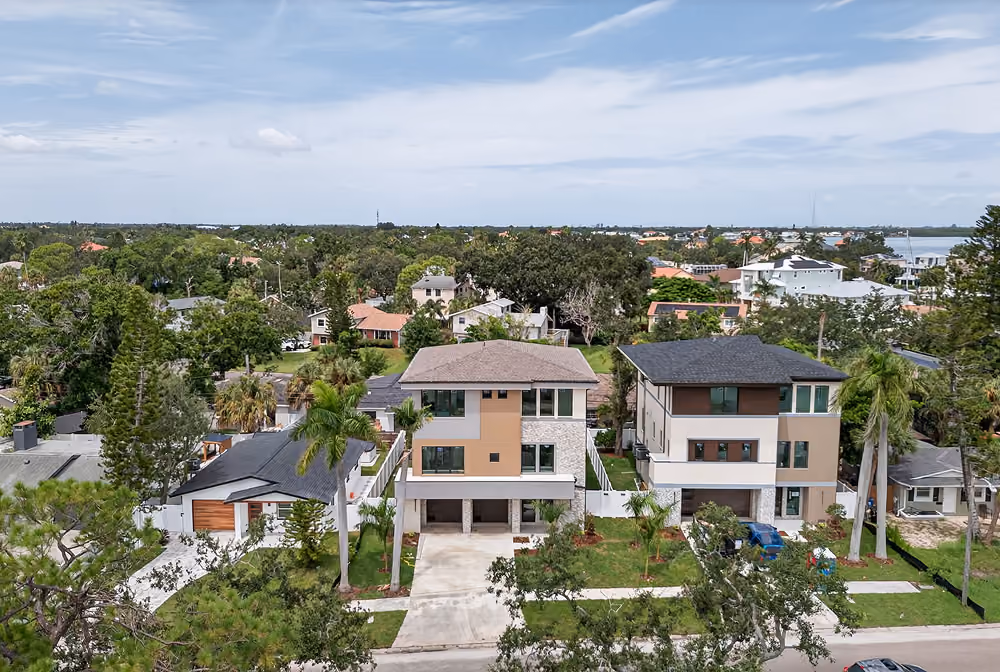 Two modern, multi-story houses with driveways and palm trees in a suburban neighborhood under a cloudy sky.
