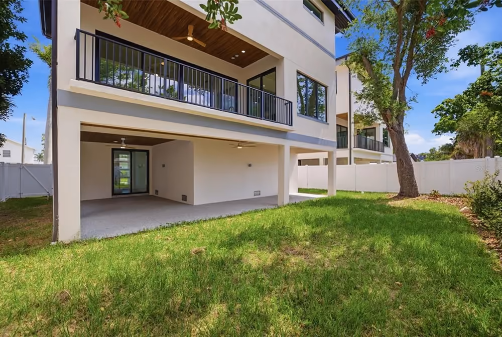 Back view of a modern two-story house with balcony and covered patio over green lawn and trees.