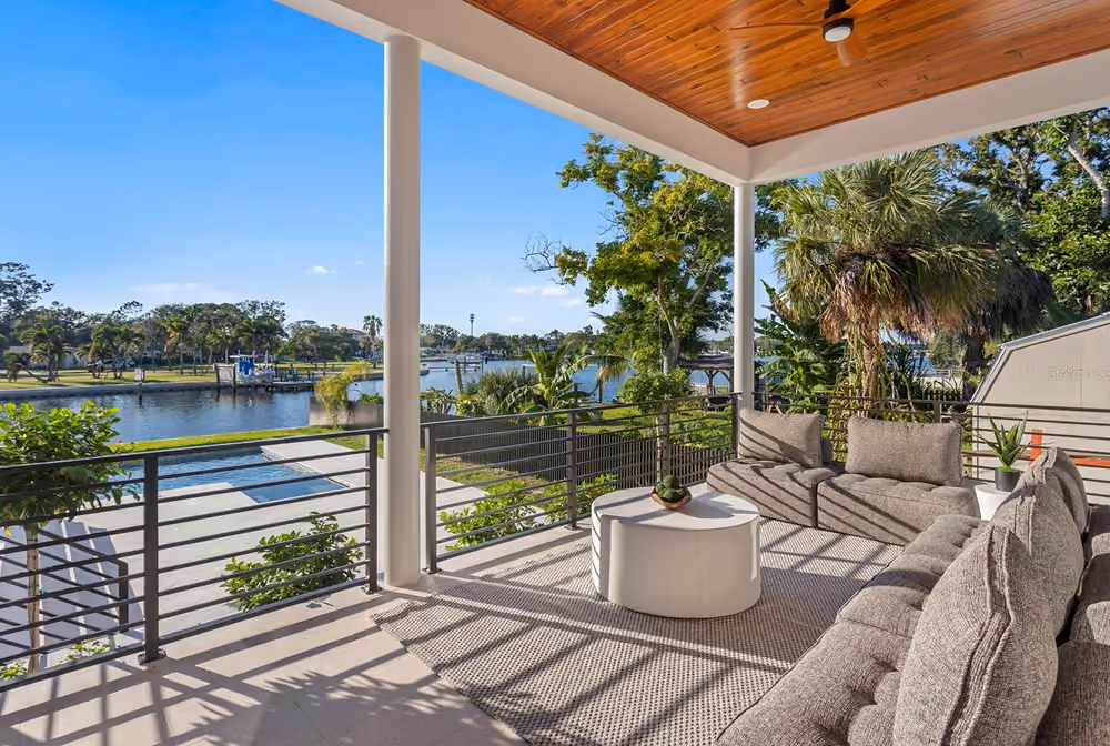 Covered patio with gray outdoor sofas, round white table, and water view with trees under clear sky.