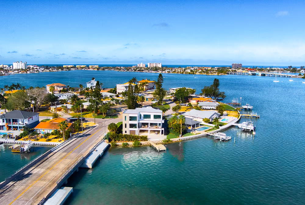 Aerial view of waterfront homes, docks, and a bridge over calm blue water in a sunny coastal neighborhood.