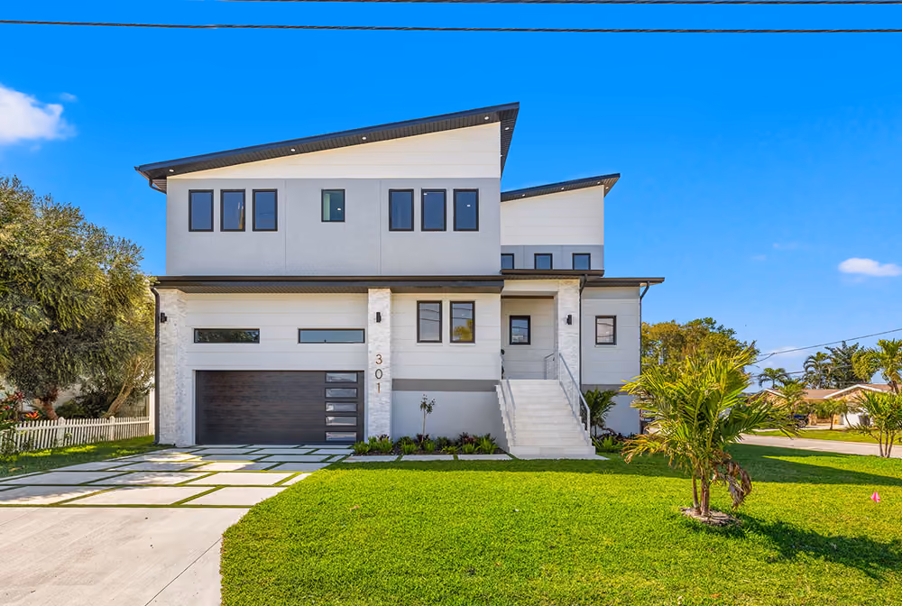 Modern two-story house with gray siding, dark garage door, stairs, and green lawn under blue sky.