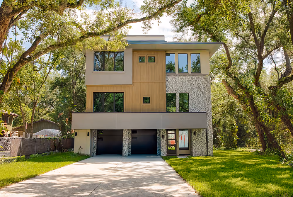 Modern three-story house with wood and stone facade, large windows, and a driveway surrounded by trees.