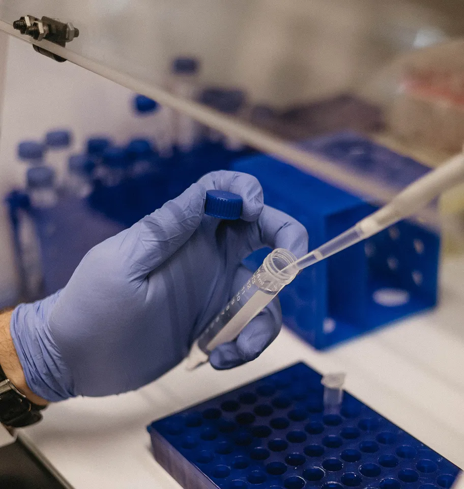 Gloved hand holding a capped tube while using a pipette to transfer liquid in a lab.