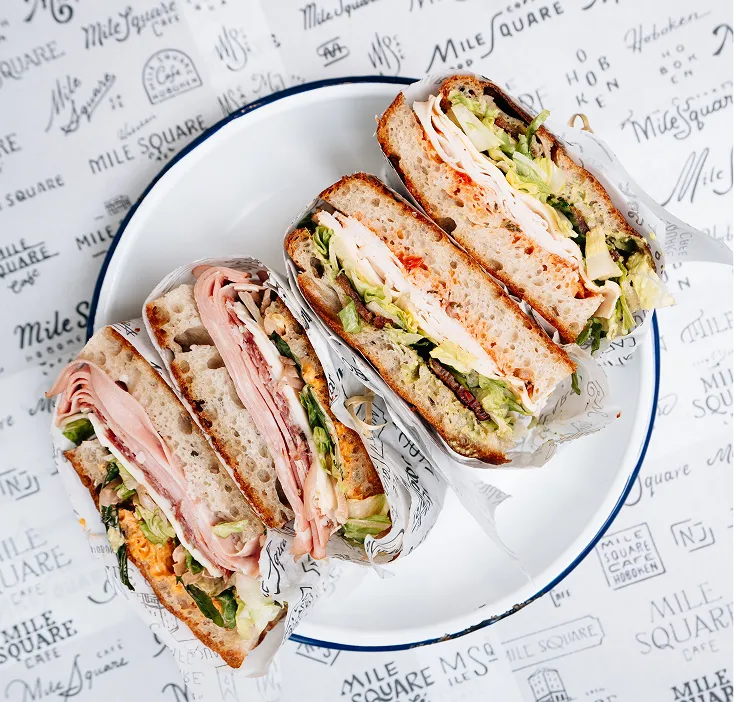 Two sandwiches on a white plate with leafy greens, deli meats, and thick crusty bread wrapped in printed paper.