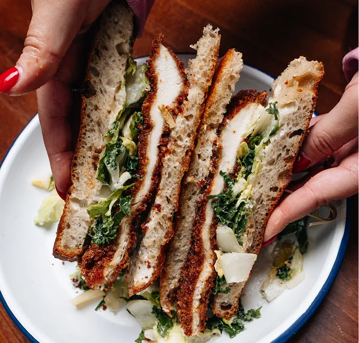 Hands holding a sandwich with breaded chicken, leafy greens, and melted cheese on a white plate with a blue rim.