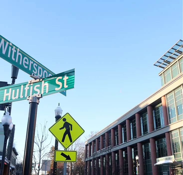 Street signs for Witherspoon St and Hulfish St above a pedestrian crossing sign next to a modern glass and brick building under a clear blue sky.
