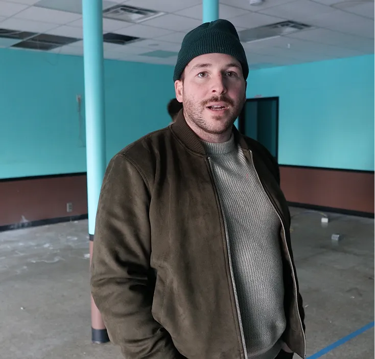 Man wearing a dark green beanie, brown jacket, and gray sweater standing in an empty room with turquoise walls and a concrete floor.