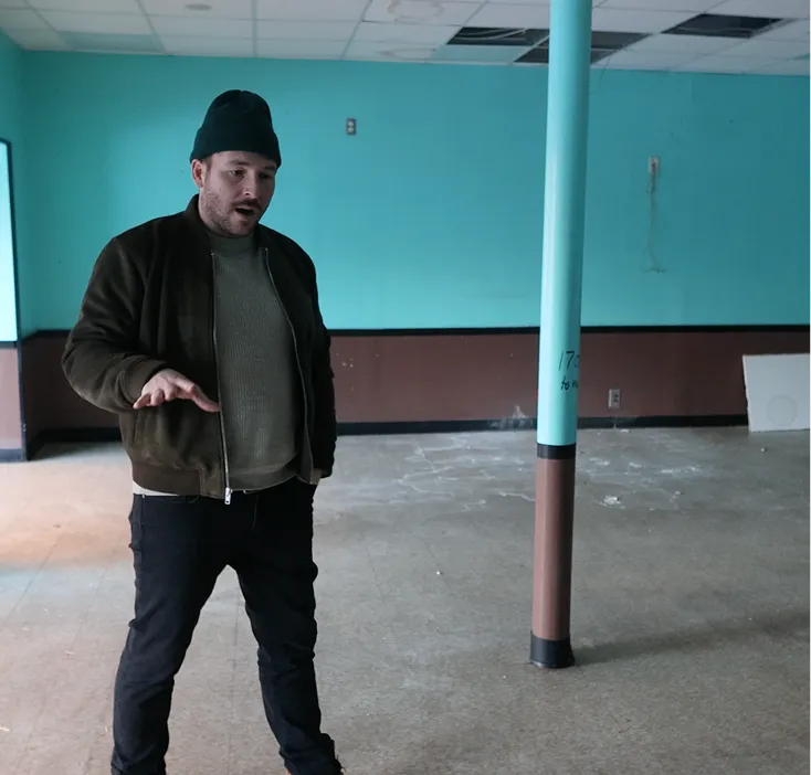Man wearing a black beanie and brown jacket stands in an empty room with turquoise walls and a turquoise support column.