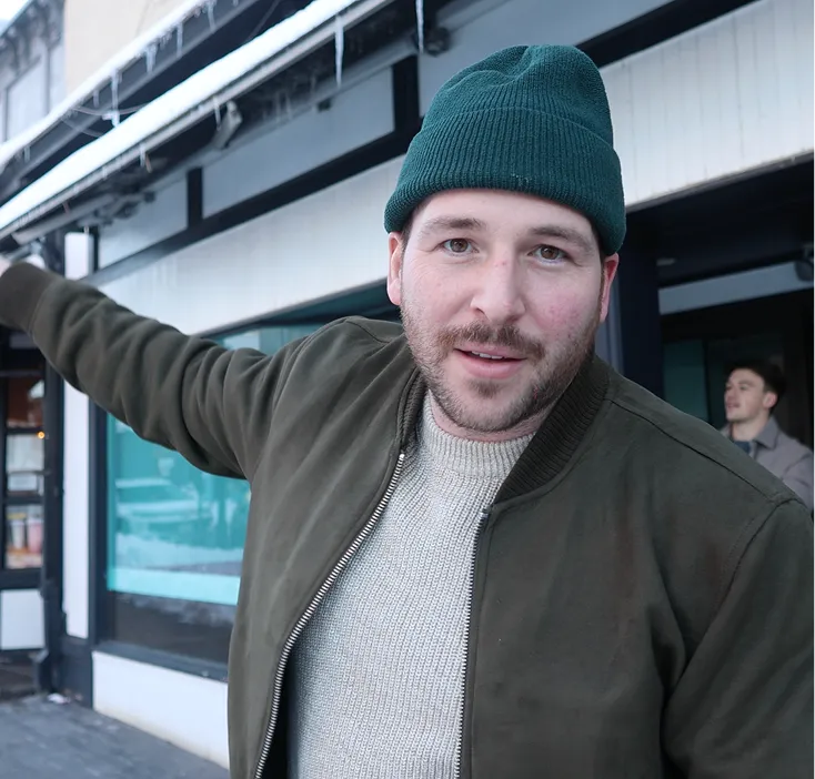 Man wearing green beanie and jacket standing outside a building with icicles hanging from the roof, gesturing with his arm extended.