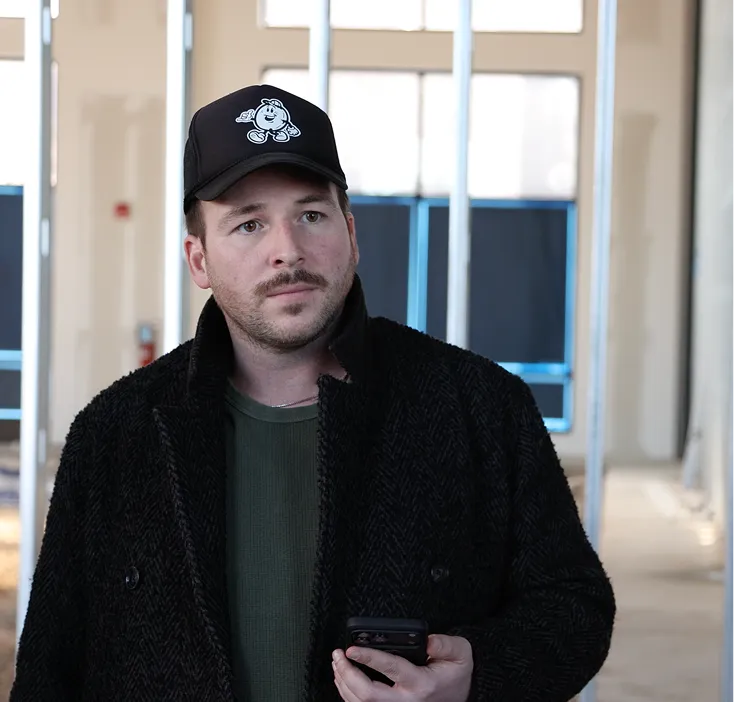 Man wearing a black baseball cap with a cartoon figure and a black jacket, holding a smartphone indoors with large windows in the background.