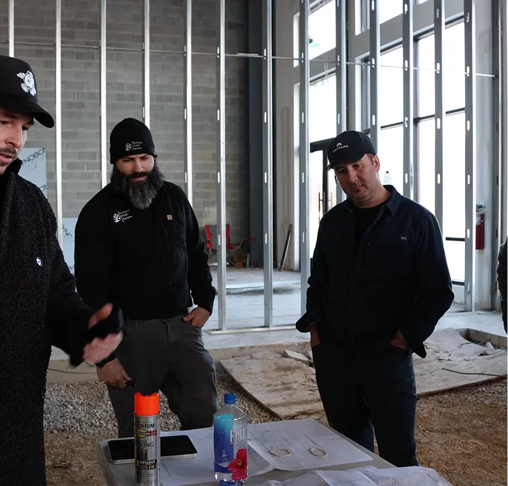 Three men wearing dark clothing and hats review construction plans on a table inside a partially built space with exposed metal framing.