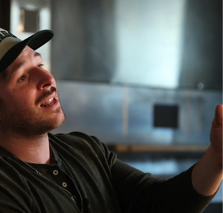 Man wearing a baseball cap and dark shirt gesturing with his hand while speaking indoors.