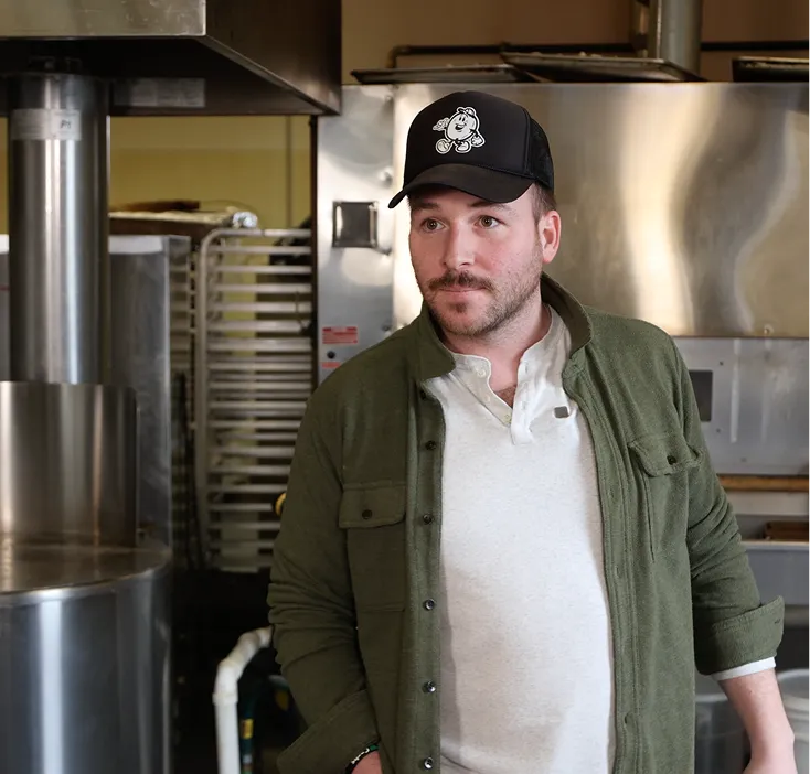 Man wearing a black baseball cap and green jacket standing in a commercial kitchen with metal equipment in the background.