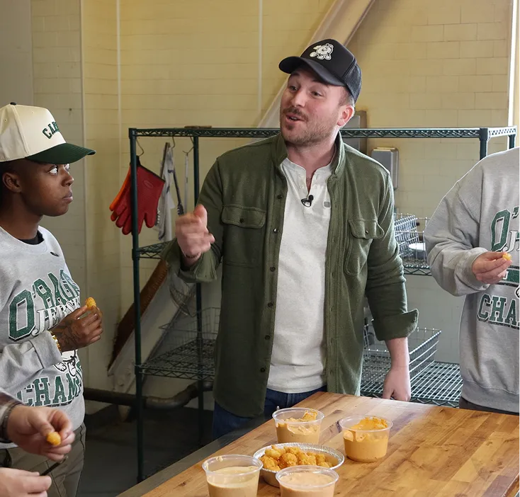 Three men tasting food around a wooden table with cups of sauce and a plate of fried snacks.