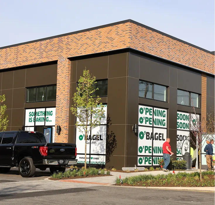 Storefront under construction with large windows and a sign saying 'Coming Soon O'Bagel' on the door, surrounded by snow and a parked silver truck.