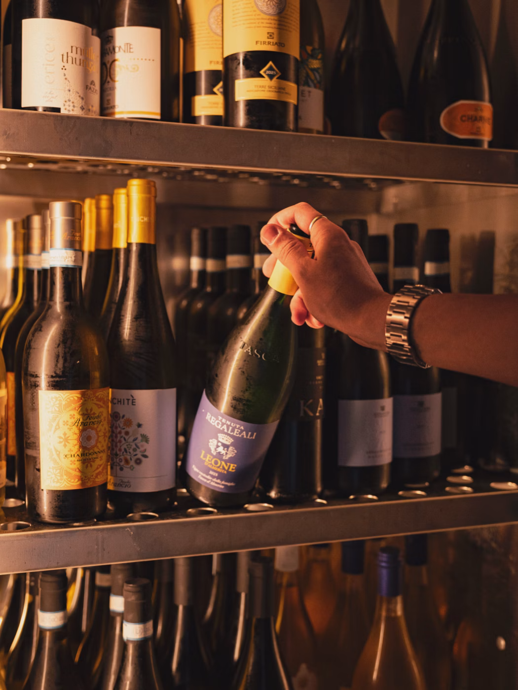 Una mano con orologio afferra una bottiglia di vino con etichetta blu in una cantina.