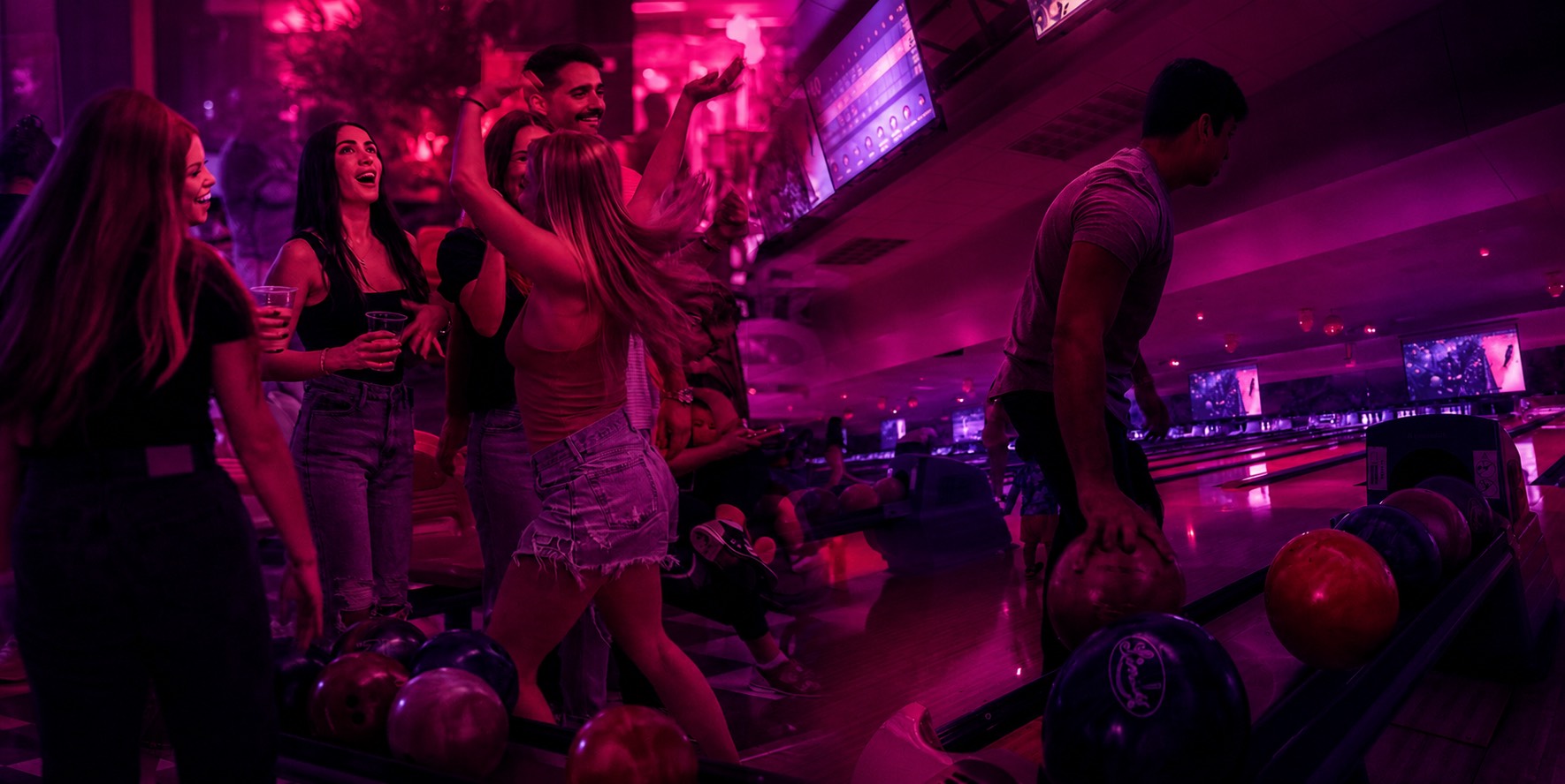 Group of young adults enjoying a night at a bowling alley with vibrant pink and purple lighting, some holding drinks and one person preparing to bowl.