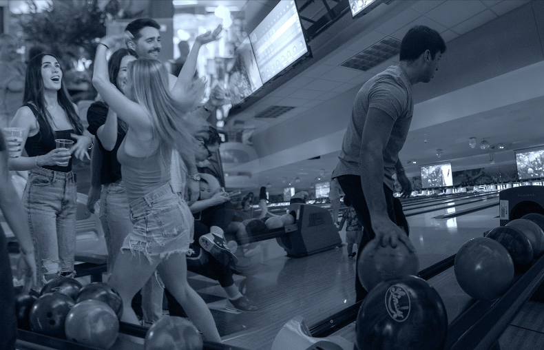 Group of young adults enjoying a game at a bowling alley, with one man preparing to bowl and others cheering and smiling nearby.