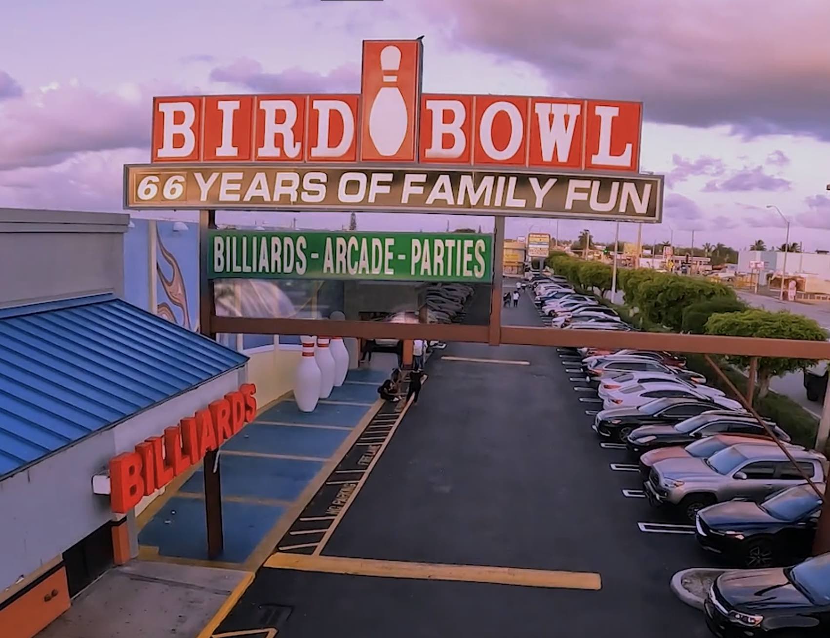 Outdoor view of Bird Bowl bowling alley with a large sign reading 'Bird Bowl 66 Years of Family Fun,' below which a green sign advertises billiards, arcade, and parties, alongside a parking lot filled with cars and blue roof building marked 'Billiards.'