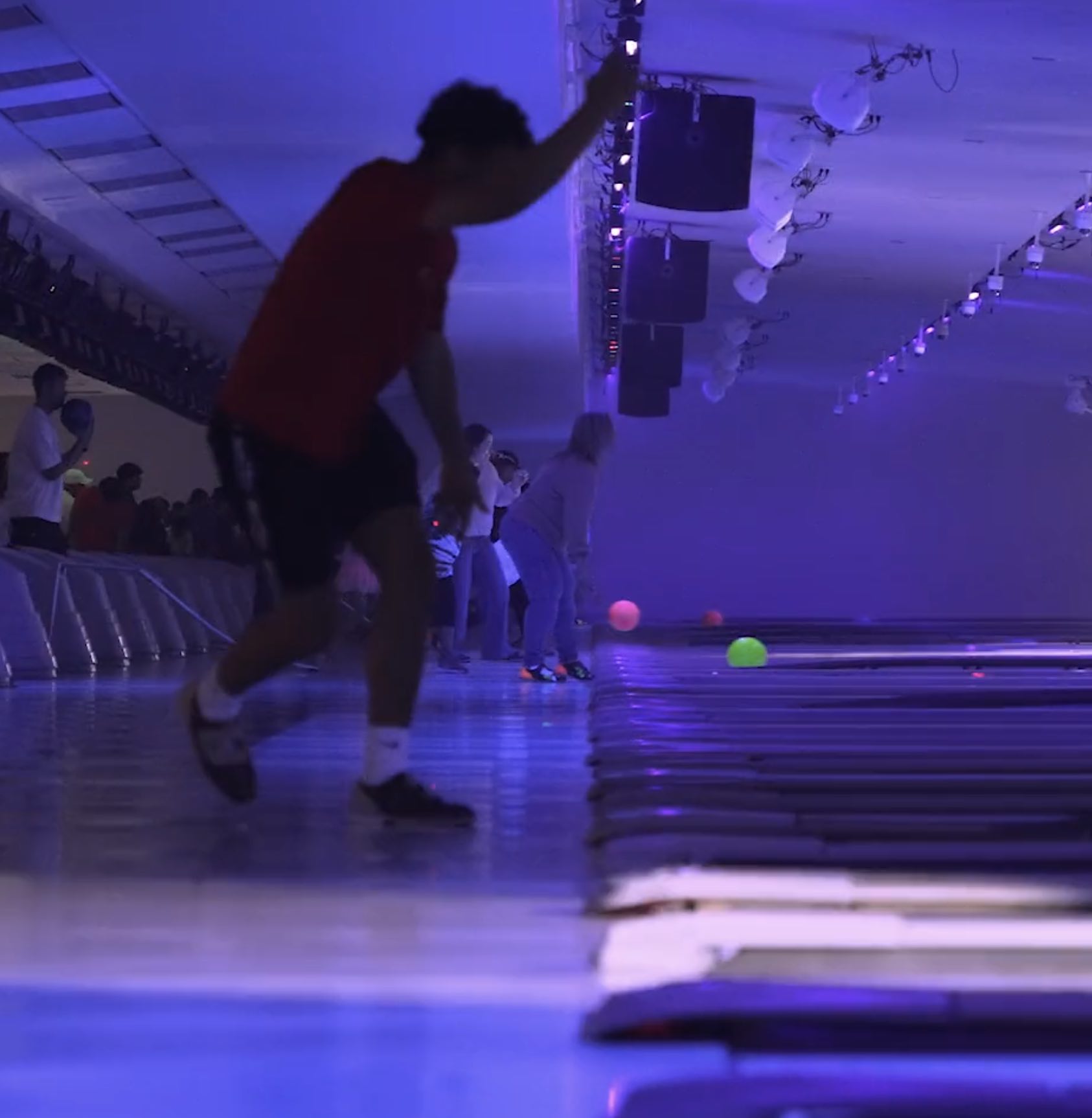 People bowling in a dimly lit alley under purple and blue lighting, with some bowling balls rolling down the lanes.