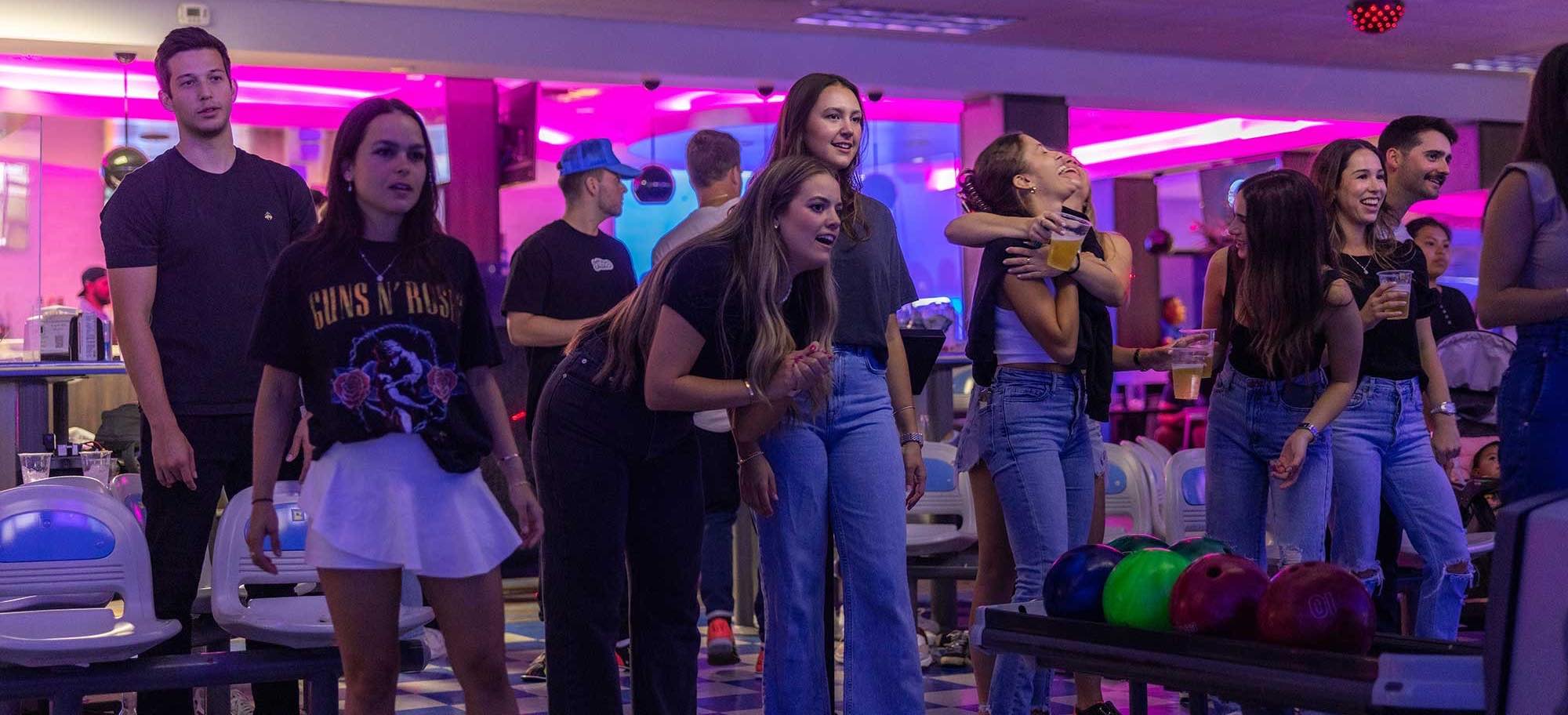 Group of young adults at a bowling alley with colorful bowling balls in the foreground, under purple neon lighting, some holding drinks and socializing.