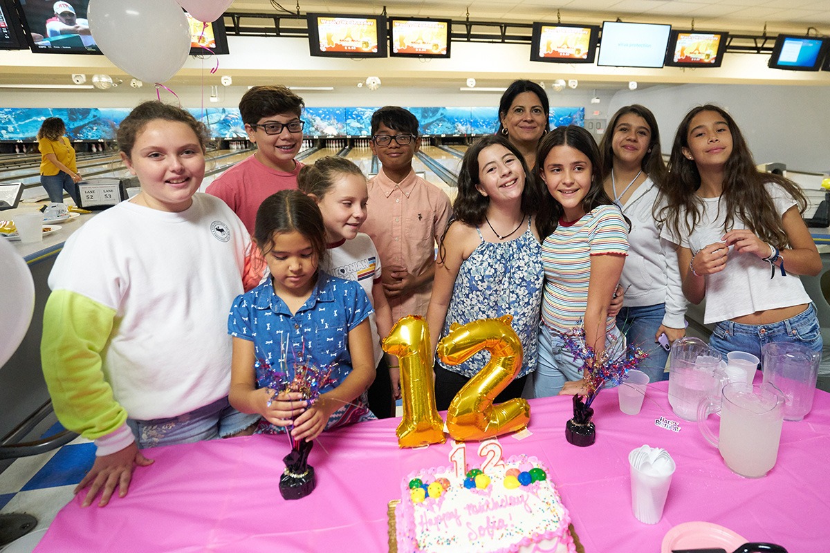 Group of children and one adult smiling and posing behind a pink table with birthday decorations, including golden balloon numbers 1 and 2, and a pink birthday cake with candles showing the number 12, at a bowling alley.