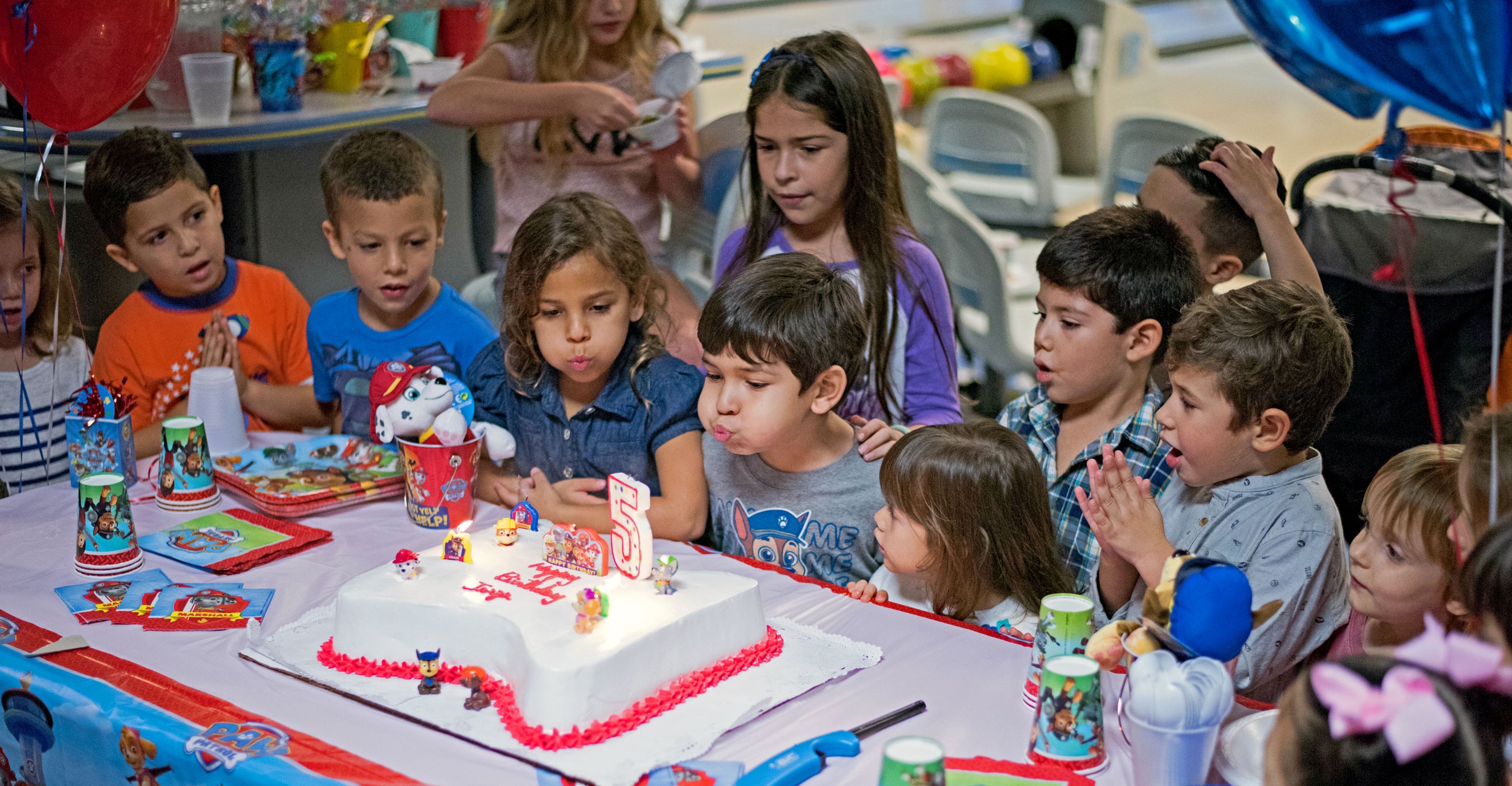Group of children gathered around a birthday cake with a large number 5 candle, as two kids blow out the candles during a party with Paw Patrol themed decorations and balloons.