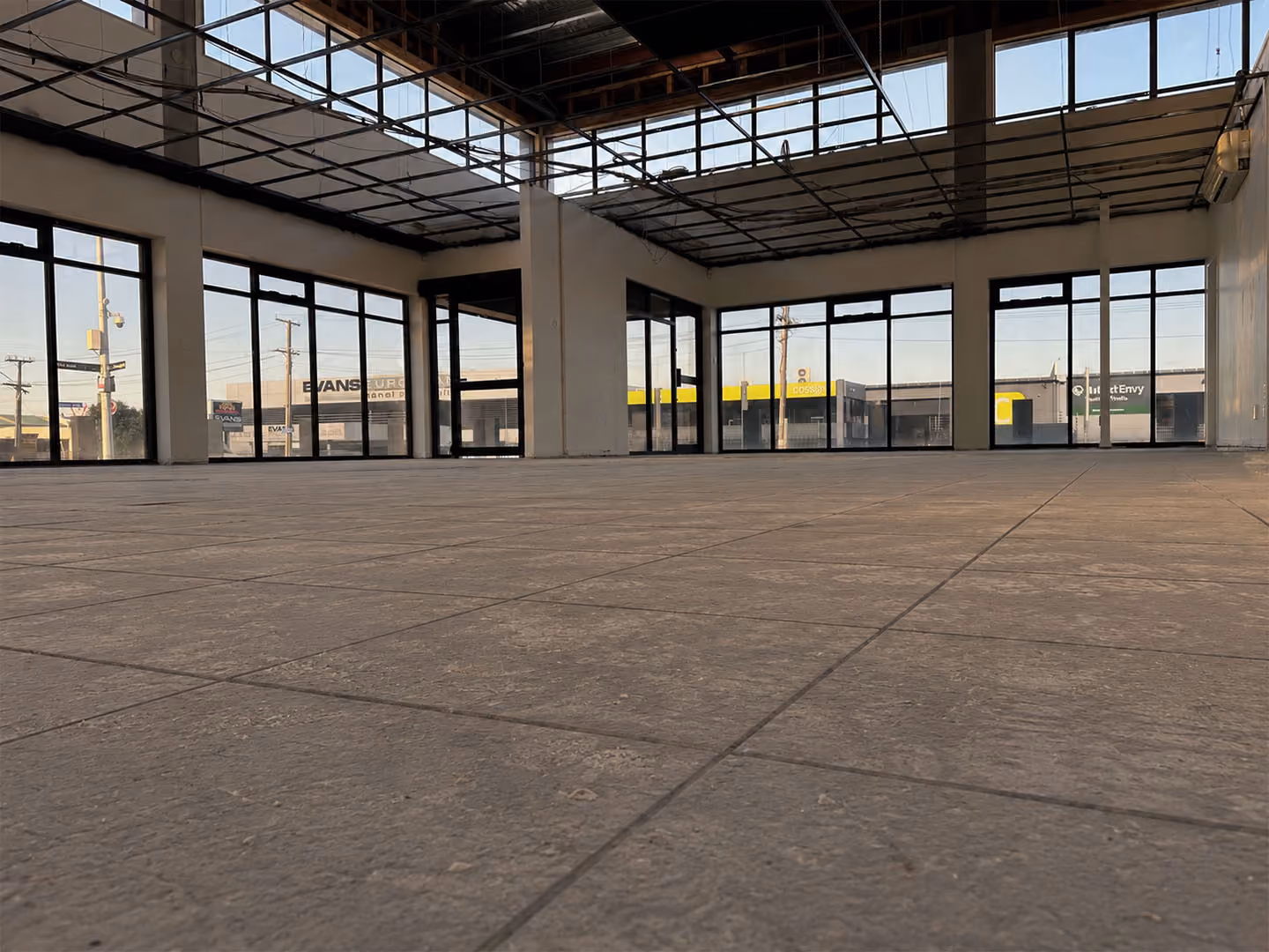 Empty retail space with large windows, tiled floor, and exposed ceiling framework.