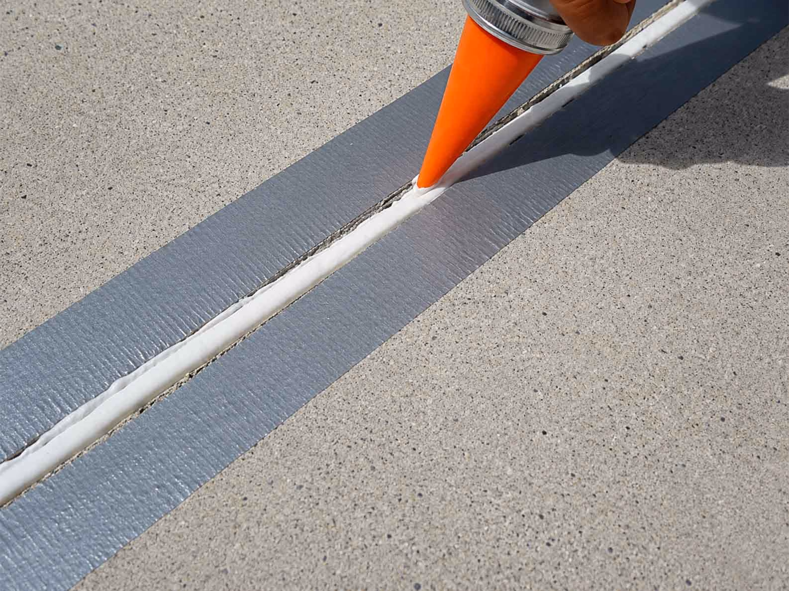 Hand applying white sealant to a concrete joint with gray tape on both sides.