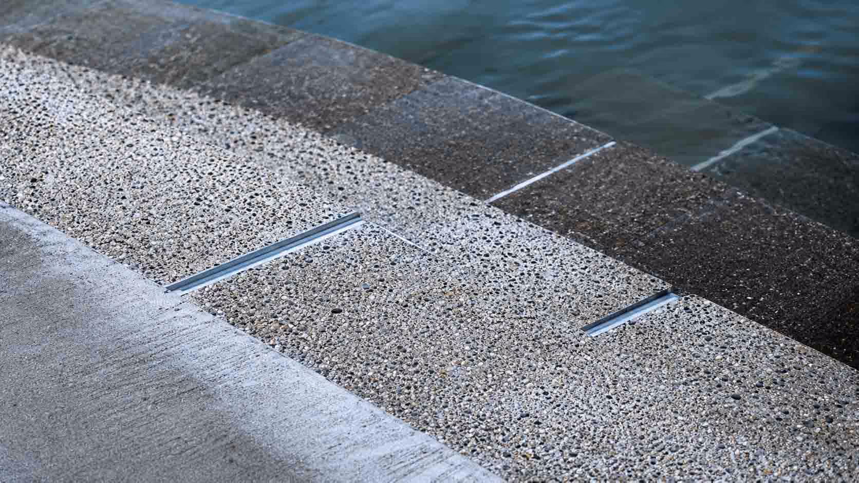 Close-up of a concrete surface with exposed aggregate and metal expansion joints near water.