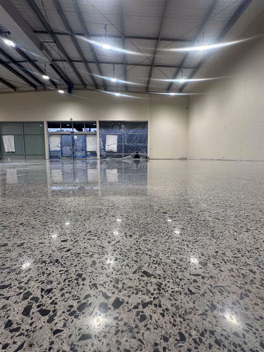 Empty commercial space with polished terrazzo floor, high ceiling, and plastic-covered windows and doors.