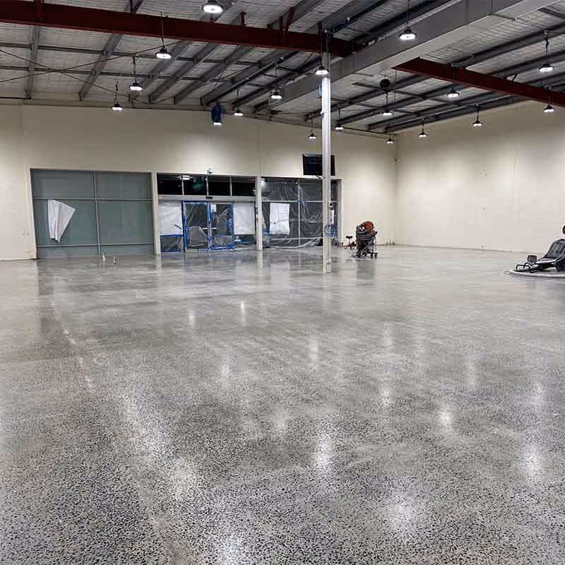 Large empty indoor space with polished concrete floor, high ceiling, and overhead lights.