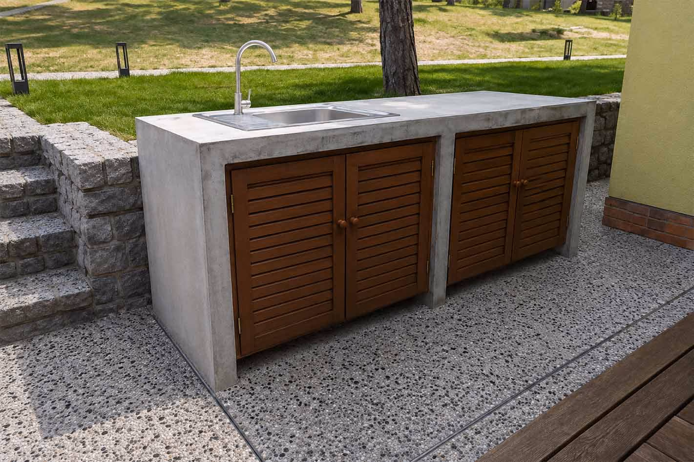 Outdoor concrete kitchen island with stainless steel sink and wooden cabinet doors on stone patio.