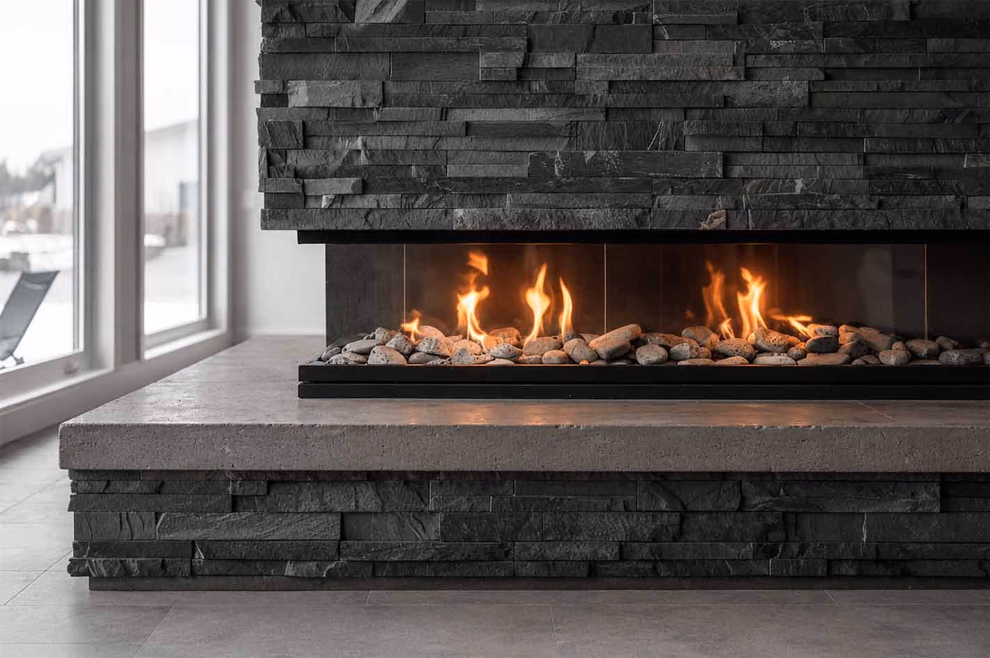 Modern gas fireplace with flames over light river rocks, surrounded by dark stacked stone and concrete hearth.
