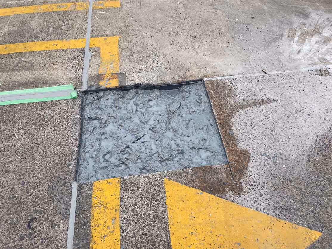 Section of concrete pavement freshly patched with wet cement surrounded by yellow road markings.