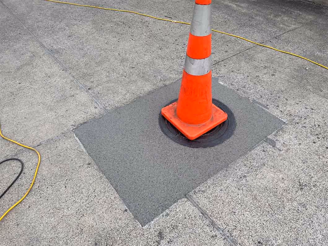 Orange traffic cone on a newly patched square section of concrete sidewalk with cords nearby.