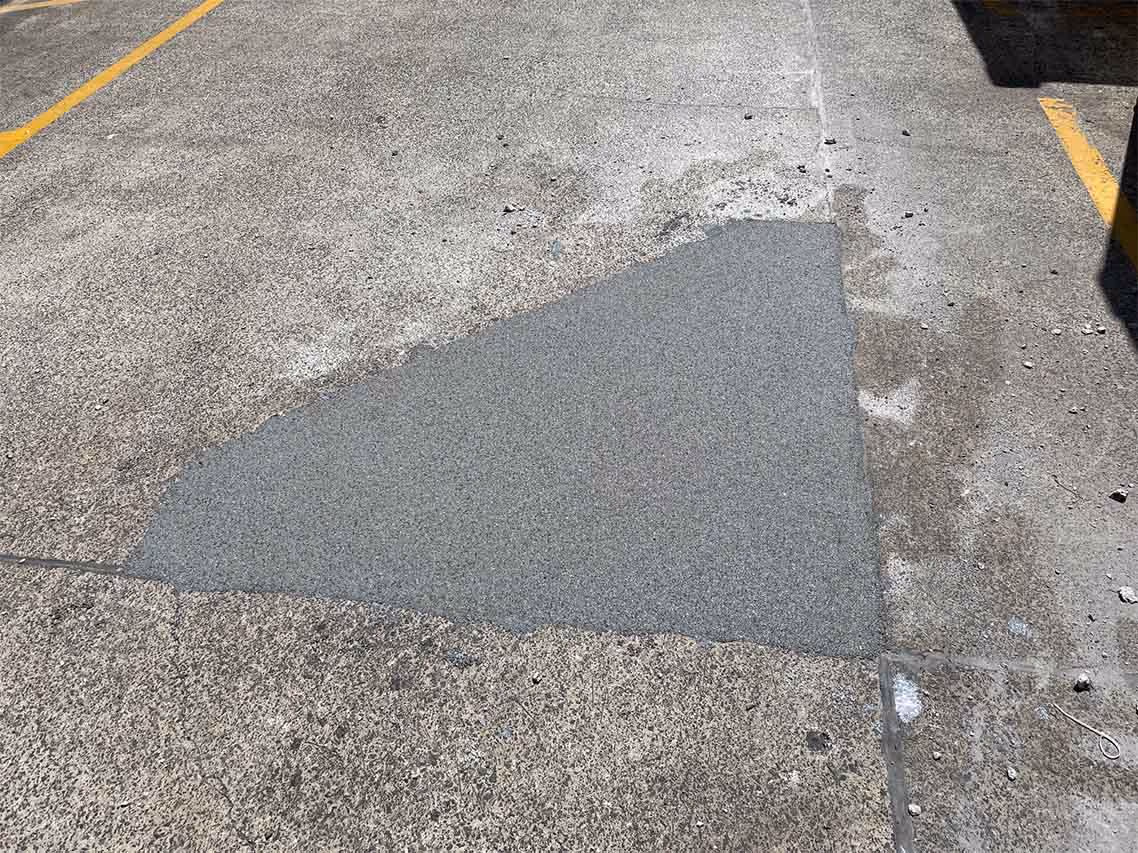 Triangular patch of fresh asphalt repair on an old concrete parking lot surface between yellow lines.