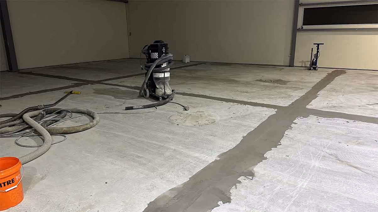 Concrete floor under repair with patching material, vacuum equipment, hoses, and orange bucket visible.