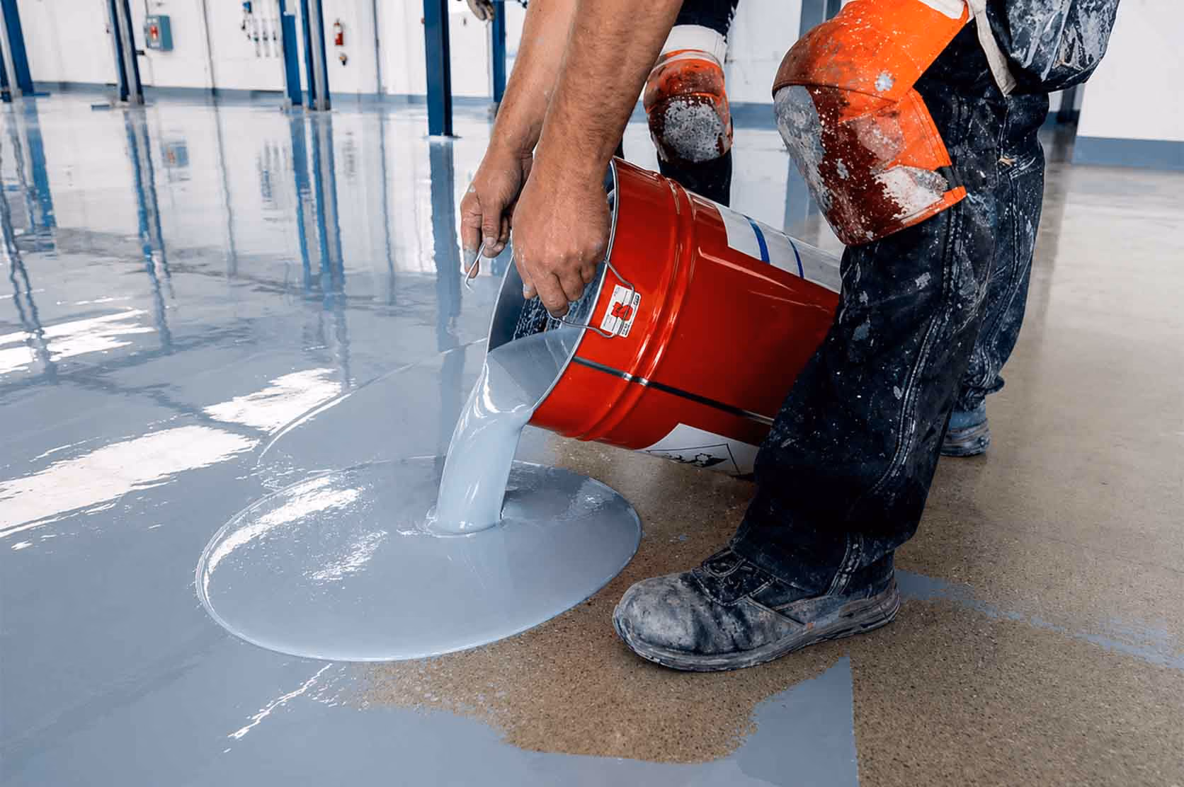 Worker pouring gray epoxy coating from a red bucket onto a concrete floor in an industrial space.