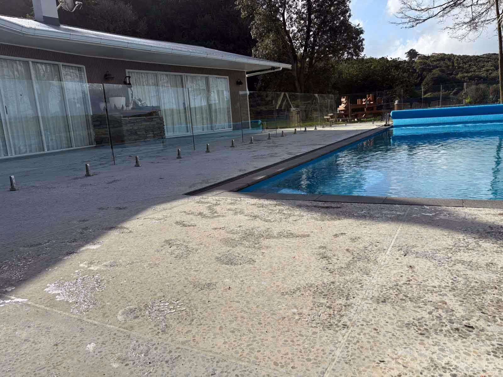 Outdoor swimming pool next to a concrete patio with glass fence panels and a house in the background.
