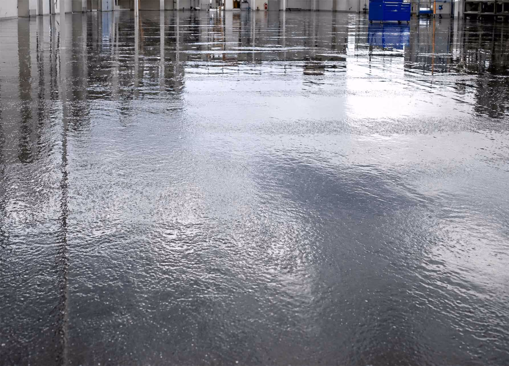 Shiny wet concrete floor reflecting lights and objects in an industrial or warehouse setting.