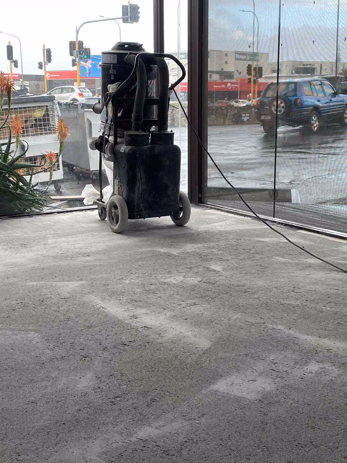 Black industrial floor cleaning machine on a concrete floor by large glass windows showing street traffic outside.