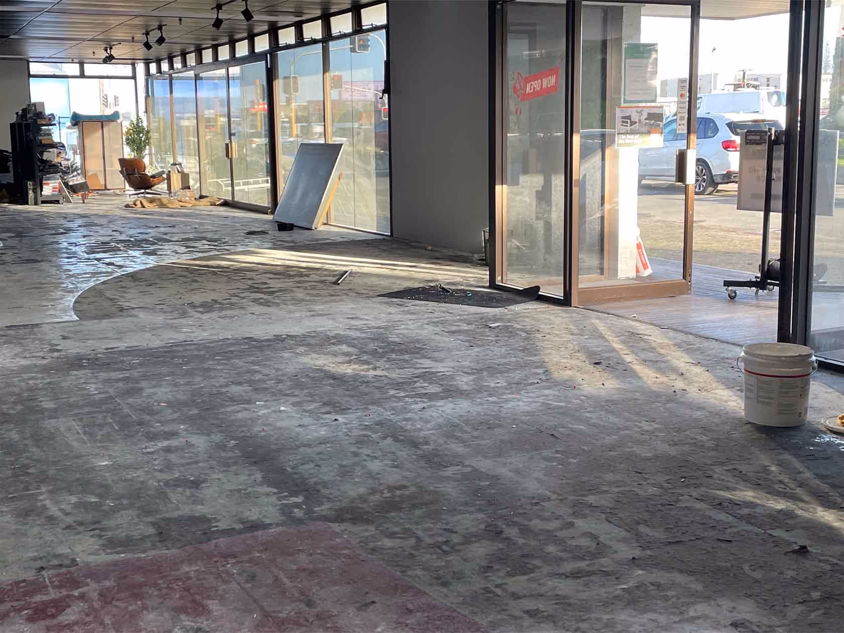 Empty commercial space with bare floor, large glass windows, open door, and scattered renovation materials.