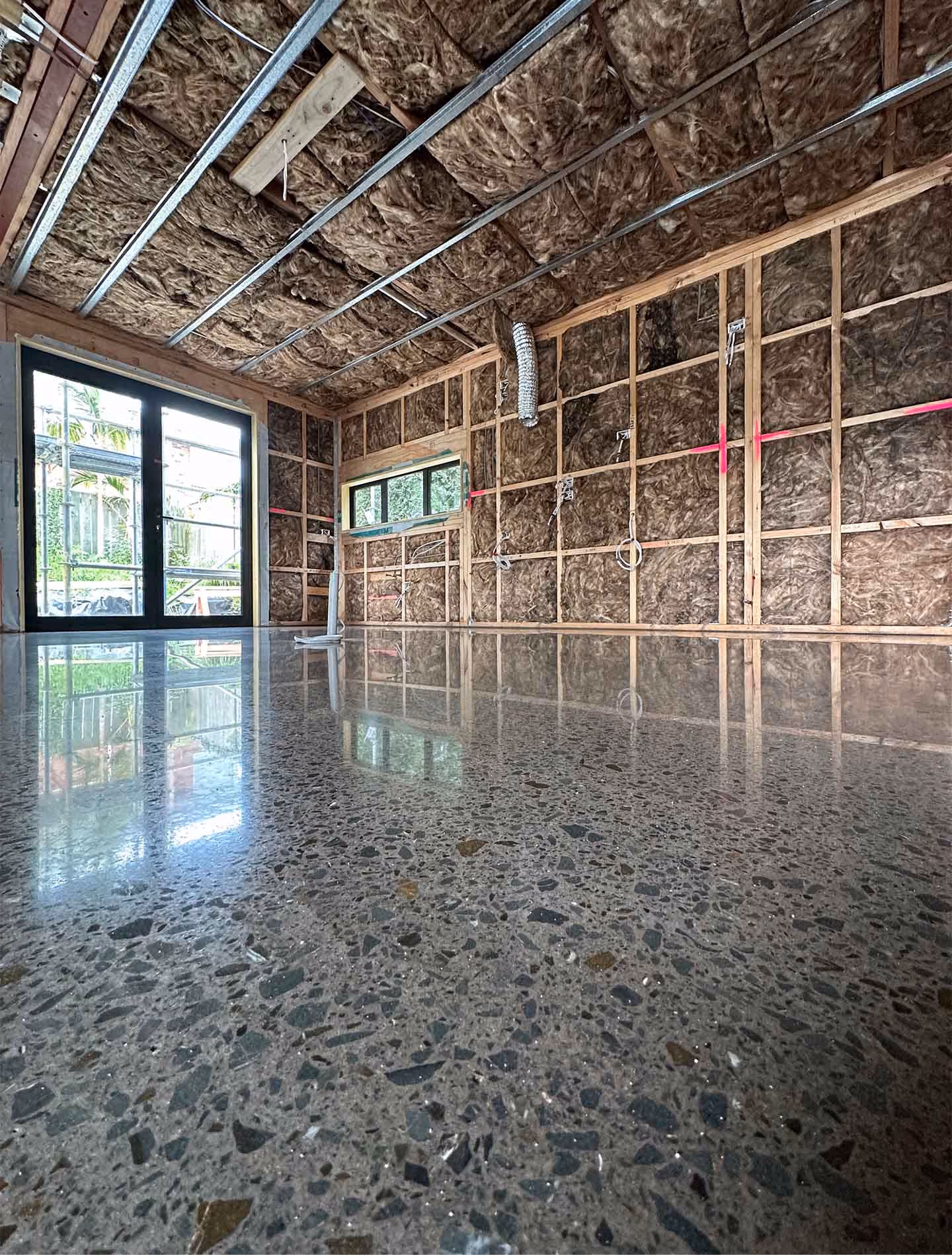Interior of a room under construction with exposed insulation and polished concrete floor reflecting windows.