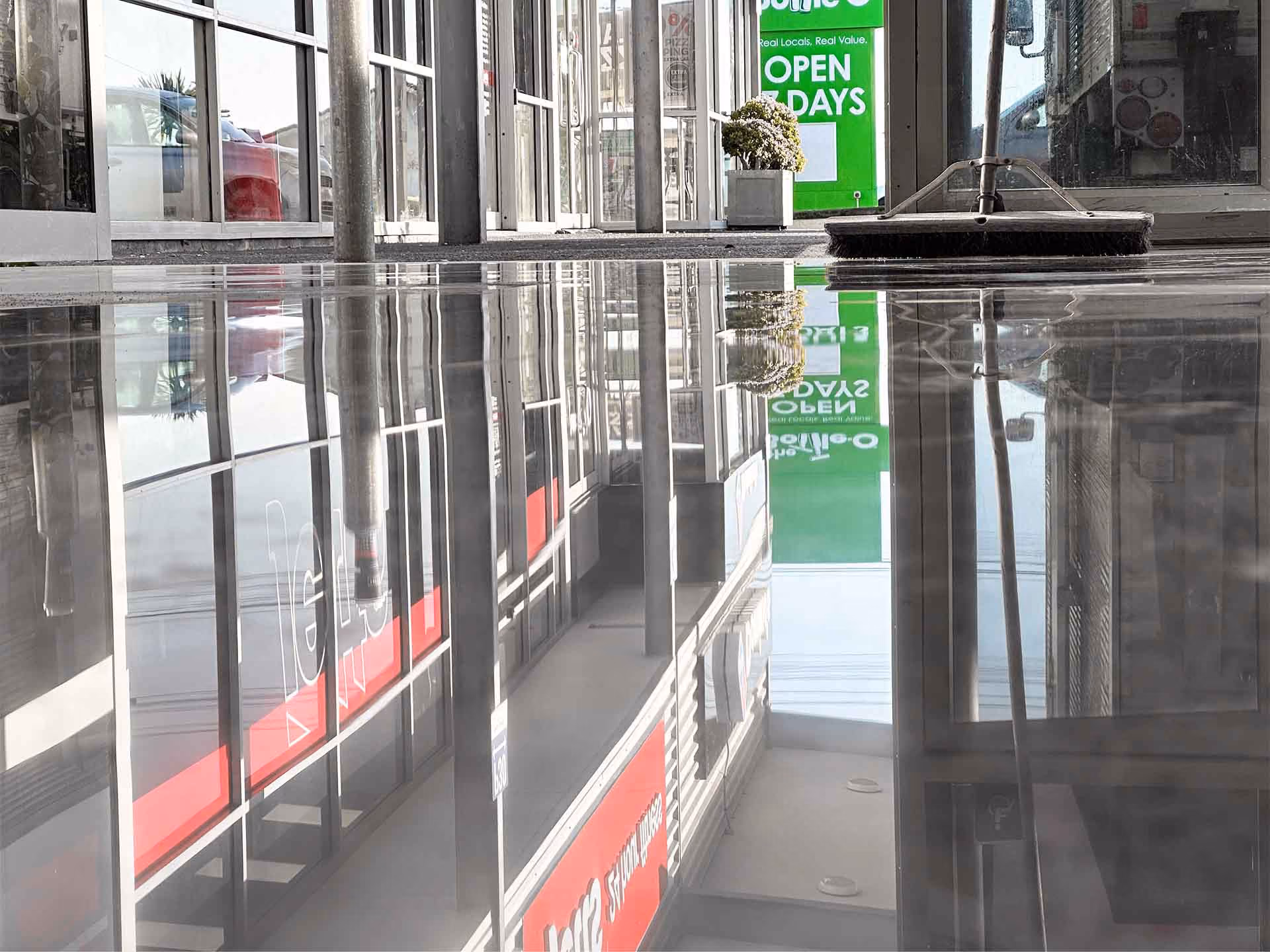 Highly polished reflective floor outside a commercial building with glass doors and a broom.