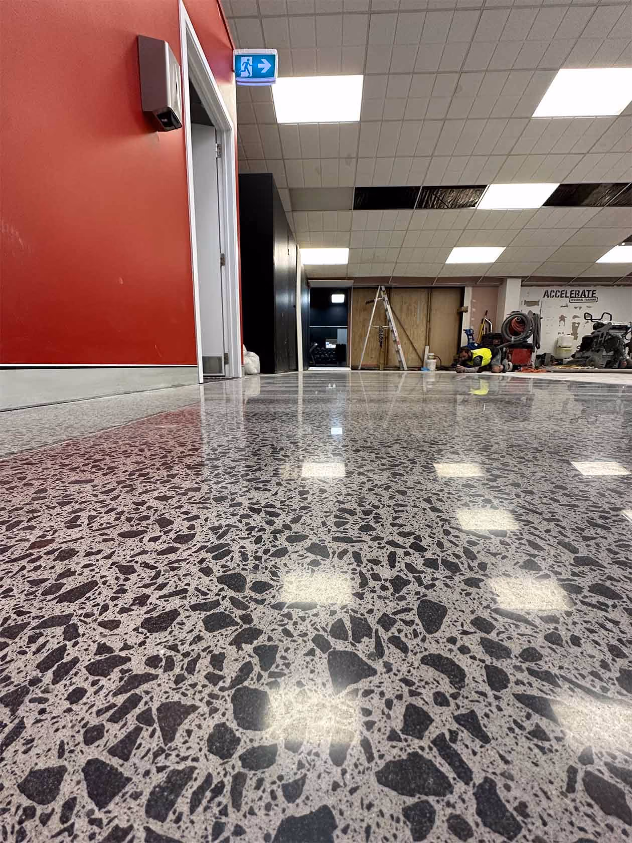 Polished terrazzo floor reflects ceiling lights in a gym with a red wall and a worker near ladder.