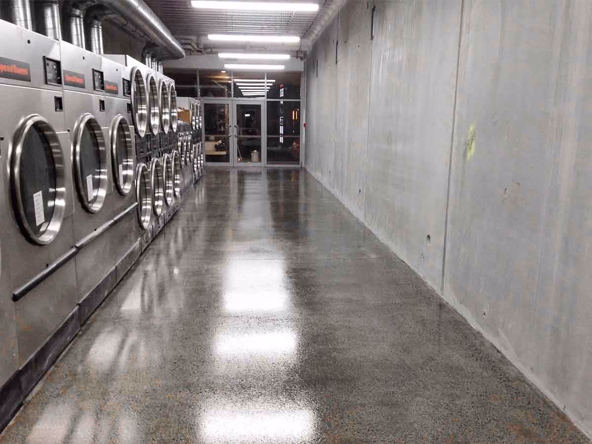Row of industrial washers or dryers along a polished concrete floor hallway in a laundromat.