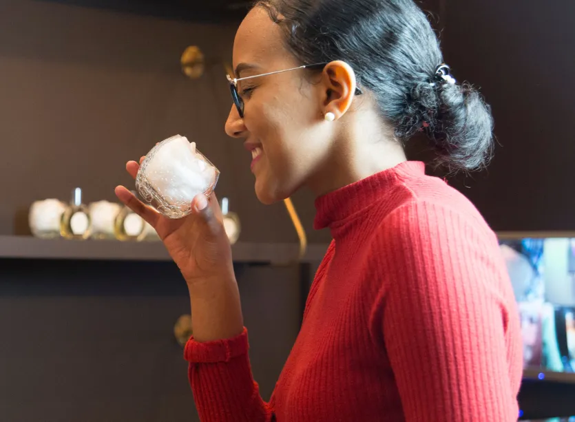 Femme souriante en pull rouge sentant une bougie dans un verre décoratif.