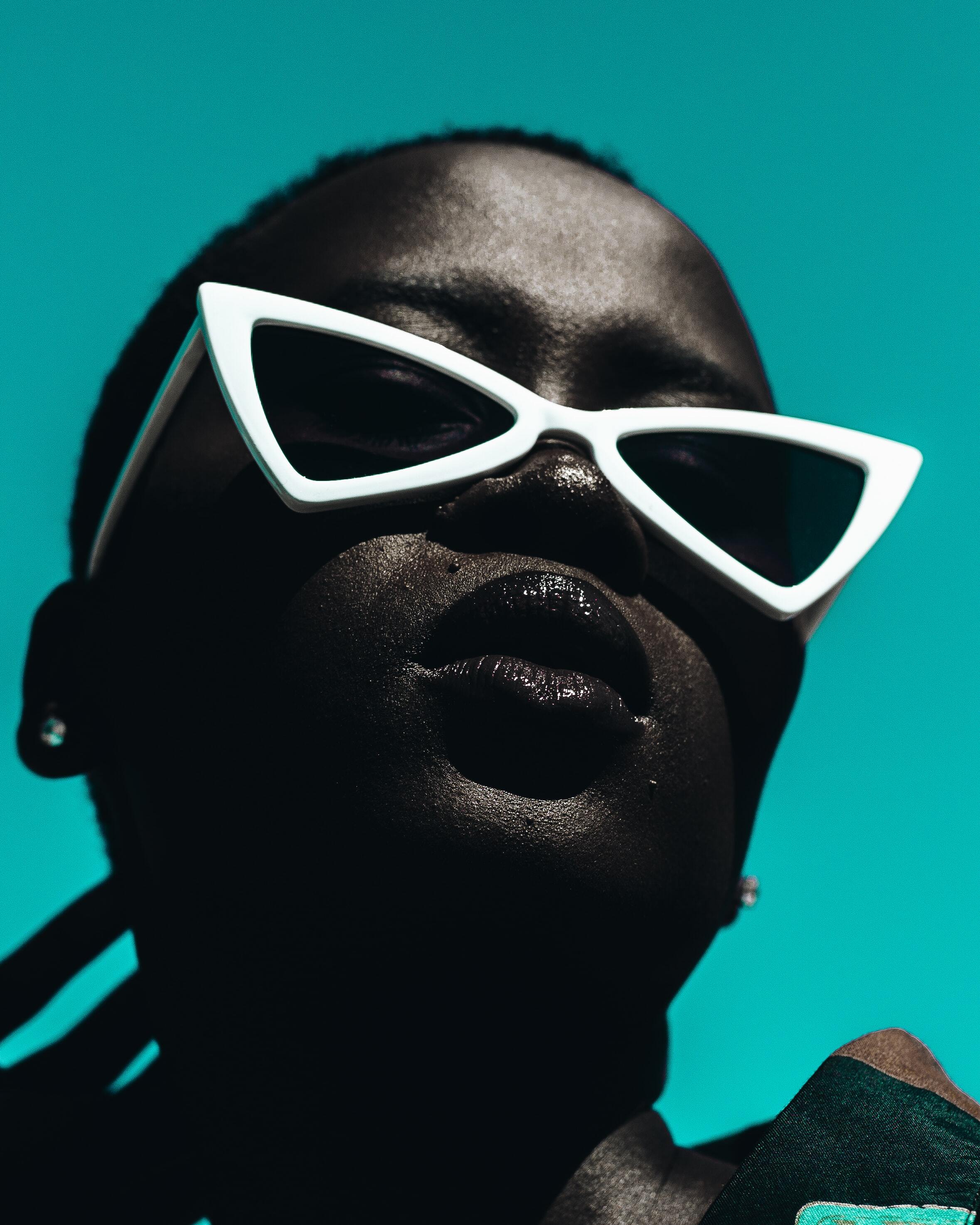 Portrait en gros plan d'une personne portant des lunettes blanches au style chat, fond turquoise vif.
