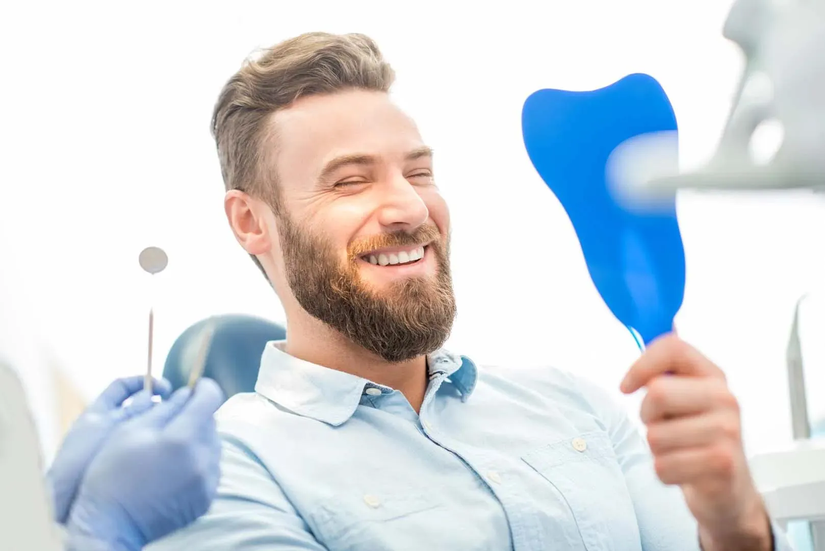 Smiling man with beard holding a blue mirror, sitting in a dentist's chair with dental tools in foreground.